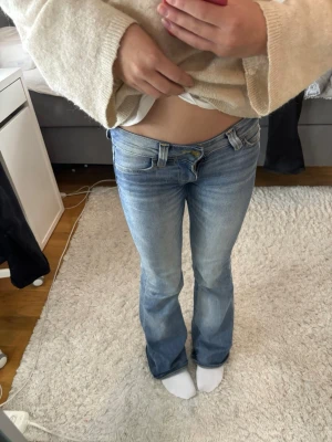 Blå Bootcut jeans från Cheap monday  - Säljer ett par ljusblå bootcut jeans med låg midja och klassiska fem fickor. Jeansen har en snygg tvätt och en patch med tryck bak i midjan. 