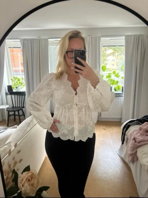 Vit broderad blus med v-ringning - Säljer en superfin vit blus med broderade blommor och v-ringning. Blusen har långa puffärmar och knappar framtill. Den har en romantisk känsla med vågig kant nertill och är gjord i ett lätt, luftigt tyg. Perfekt att styla med jeans eller kjol för en trendig look.