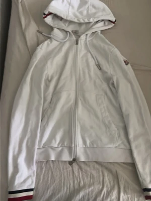 Vit hoodie med dragkedja och ränder - Snygg vit hoodie med dragkedja och huva. Har fickor framtill och coola ränder i rött, vitt och blått på huvan och ärmsluten. Perfekt för en avslappnad streetstyle-look. Materialet känns mjukt och bekvämt, och passformen är klassisk med långa ärmar.