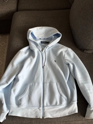 Ljusblå hoodie från Polo Ralph Lauren - Snygg ljusblå hoodie från Polo Ralph Lauren med dragkedja framtill och två stora fickor. Klassisk broderad logga på bröstet och mjukt bomullsmaterial. Perfekt för en chill och stilren look.