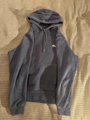 Mörkblå hoodie från Lacoste - Snygg mörkblå hoodie från Lacoste med klassisk krokodillogga på bröstet. Hoodien har en stor ficka framtill, dragsko i huvan och ribbade muddar vid ärmar och nederkant. Perfekt för en chill och avslappnad stil.