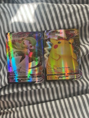 Pokémonkort Gardevoir VMAX & Pikachu - Två glittriga Pokémonkort med motiv av Gardevoir VMAX och Pikachu VMAX. Korten har färgstarka illustrationer och holografisk yta som reflekterar ljus. Perfekt för dig som gillar att samla eller spela Pokémonkort. Tillverkade i laminerad papp.