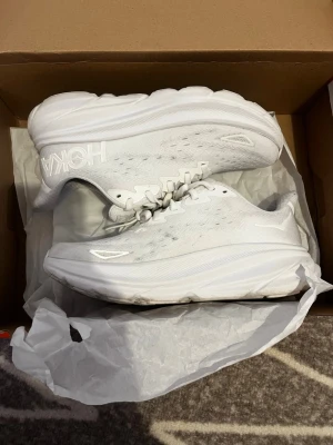 Vita Hoka sneakers  - Säljer ett par helvita sneakers från Hoka med riktigt chunky sula och ventilerande mesh-ovandel. Skorna har klassisk snörning och diskret logga på sidan. Perfekta för dig som gillar sportig och clean stil. Extremt sköna och endast använda 1 gång. Säljs pga de inte används. Pris kan diskuteras😊