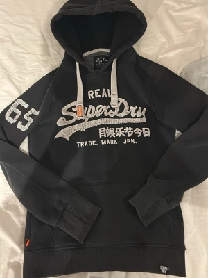 Supedry hoodie  - Säljer en svart hoodie från Superdry med stort vitt tryck på bröstet och siffran 65 på ärmen. Hoodien har vita dragsnören, känguruficka och ribbade muddar. Insidan är mjuk och borstad för extra komfort. Perfekt för en avslappnad streetwear-look.