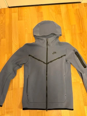 Blå Nike hoodie med dragkedja XS - Snygg blå hoodie från Nike med hel dragkedja framtill och svarta detaljer. Har huva, två fickor fram och en extra ficka med dragkedja på ärmen. Tillverkad i mjukt material som känns skönt mot huden. Perfekt för en sportig och avslappnad stil.