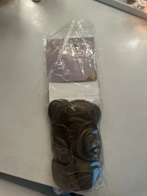Brun lockig hårförlängning clip-on - Säljer en brun hårförlängning med mjuka lockar som enkelt fästs med clips. Perfekt för dig som vill ha längre och fylligare hår på nolltid. Färgen är en naturlig brun nyans med lite skiftningar för ett mer levande intryck. Materialet är syntetiskt och känns mjukt.