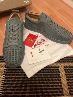 Blåa gråa Louboutin sneakers med nitar - Snygga grå sneakers från Christian Louboutin i mocka med matchande grå sula. Skorna har coola nitar framtill och klassisk låg siluett. Perfekta för dig som vill sticka ut med en exklusiv och edgy stil. Levereras med dustbag och äkthetskort.