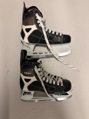 Svarta CCM skridskor med vita detaljer - Säljer ett par klassiska svarta CCM skridskor med vita detaljer och vita snören. Skridskorna har robust konstruktion, högt skaft och silverfärgade skenor. Perfekta för hockey eller allmän åkning på isen. Lite slitna fram kolla andra bilden 