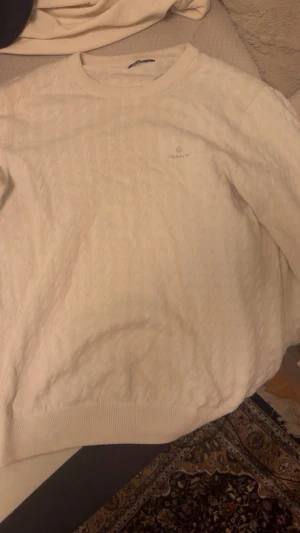 Vit kabelstickad tröja från Gant - Snygg vit kabelstickad tröja från Gant med rund halsringning och diskret logga broderad på bröstet. Tröjan har ribbade muddar vid ärmslut och nederkant, perfekt för lager på lager och en clean look.