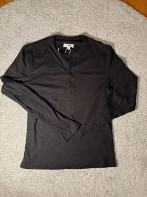 Svart långärmad topp från COS - Säljer en svart långärmad topp från COS med rund halsringning och knappar framtill. Enkel och stilren design som passar till mycket. Perfekt basplagg i garderoben.