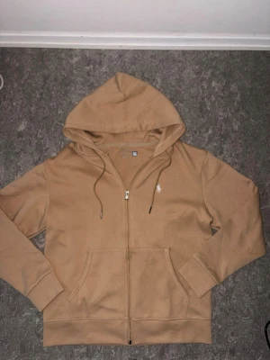Beige hoodie med dragkedja - Snygg beige hoodie med dragkedja framtill och två stora fickor. Har huva med dragsko och ribbade muddar vid ärmar och nederkant. Liten vit logga broderad på bröstet. Perfekt för en chill och avslappnad stil.