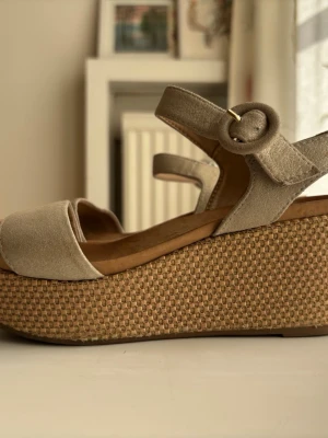 Unisa beige platåsandaler med flätad sula storlek 36 - Snygga beige sandaler med breda remmar och justerbart spänne i mocka från Unisa. Platåsulan är flätad i naturmaterial och ger extra höjd. Storlek 36, från Unisa