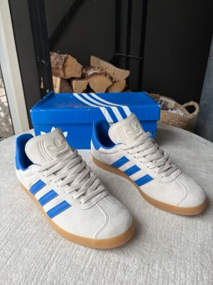 Adidas Gazelle - Saljer nu dessa riktigt fina Adidas Gazelle skorna i färgen Beige med gummi färgade sulor och blåa detaljer. Storlek - 42 Skick - 10/10 Försöker skicka samma dag annars så snabbt jag kan! Hör av dig vid fler frågor!