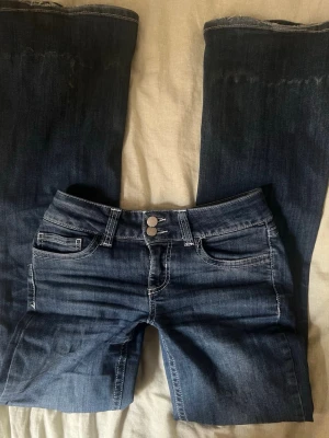 Mörkblå bootcut jeans med dubbla knappar - Snygga mörkblå bootcut jeans med dubbla silverknappar i midjan och kontrastsömmar. Klassisk femficksmodell med bälteshällor och lätt slitning på låren. Tillverkade i stretchigt denimtyg för extra komfort.