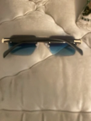 Svarta solglasögon med blå glas - Säljer ett par trendiga solglasögon med rektangulära, blåtonade glas och svarta bågar. Snygga gulddetaljer vid gångjärnen ger en exklusiv känsla. Perfekta för dig som vill sticka ut med en modern accessoar.