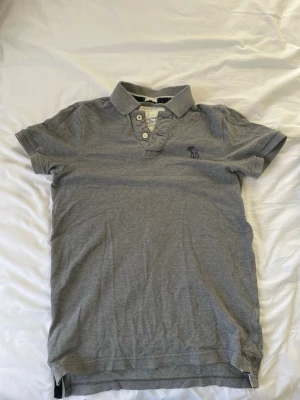 Grå pikétröja från Abercrombie & Fitch - Snygg grå pikétröja från Abercrombie & Fitch med klassisk krage och tre knappar framtill. Tröjan har korta ärmar och en diskret logga broderad på bröstet. Perfekt till jeans eller shorts för en avslappnad look. Om ni har funderingar fråga gärna.