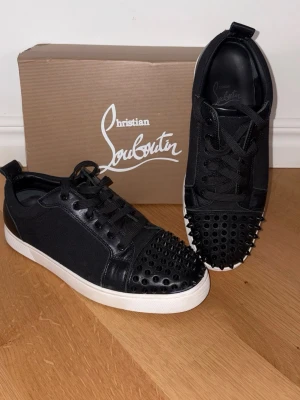 Christian Louboutin svarta sneakers - Svarta sneakers från Christian Louboutin med vita sulor och coola svarta nitar på tån. Skorna har en mix av läder och textil, svarta skosnören och en klassisk låg siluett. Perfekta för dig som vill sticka ut med en edgy och lyxig stil.