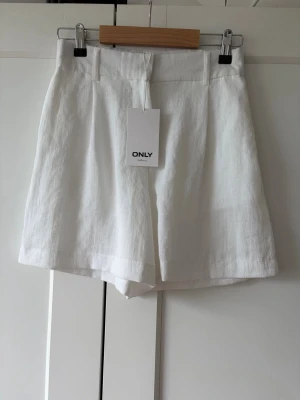 Vita linneshorts från ONLY - Helt nya, Snygga vita shorts från ONLY i luftigt linne. De har hög midja, breda benslut och hällor för bälte. Perfekta för varma dagar och ger en clean och fräsch look. Stängs med dragkedja och knapp framtill.
