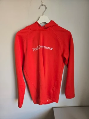 Hoodie från Peak Performance - Säljer en snygg orange  hoodie från Peak Performance med vit logga på ryggen och liten logga på huvan. Hoodien har långa ärmar, huva och är tillverkad i mjukt material. Perfekt för en sportig och avslappnad stil.