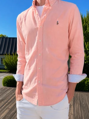 Korralrosa/Ljusrosa skjorta från Ralph Lauren - Säljer en ljusrosa skjorta från Ralph Lauren med klassisk button-down krage och broderad logga på bröstet. Skjortan har långa ärmar med randiga manschetter och vita knappar framtill. Perfekt för dig som gillar stilrena plagg med en preppy touch.