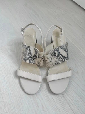 Vagabond- Beige klackar - Snygga sandaler från Vagabond i beige med bred rem i ormmönstrat material och justerbar rem runt ankeln. Skorna har en stabil klack i trälook och öppen tå. Sköna att gå i länge
