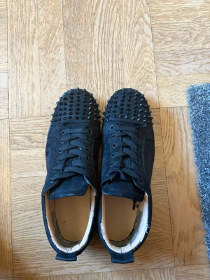 Svarta sneakers med nitar och röd sula - Svarta loubs  med  svarta nitar på tån och ovandelen. Skorna har snörning och en ikonisk röd sula undertill. Materialet  mocka och textil. stil. Äktehtesbevis privat