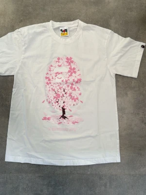 Vit BAPE t-shirt med rosa tryck - Vit t-shirt från BAPE med ett stort rosa tryck av ett träd med blomblad som formar det klassiska BAPE-ansiktet på framsidan. T-shirten har rund hals och korta ärmar. Perfekt för dig som gillar streetwear och unika prints.
