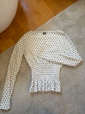 Vit prickig blus från Gina Tricot - Supersöt vit blus från Gina Tricot med svarta prickar och smockad midja som ger en snygg siluett. Blusen har långa ärmar och lätt volang nedtill. Perfekt för dig som gillar trendiga och feminina detaljer.