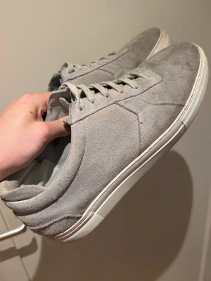 Gråa sneakers i mocka från Axel Arigato - Snygga gråa sneakers från Axel Arigato i mjuk mocka med vita sulor. Klassisk låg modell med stilrena sömmar och matchande grå skosnören. Perfekta för dig som gillar minimalistisk och clean stil. Storlek 47