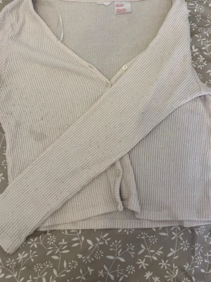 Beige ribbad kofta med knappar - Säljer en beige ribbad kofta med v-ringning och långa ärmar. Koftan har en croppad passform och stängs med vita knappar framtill. Perfekt att slänga över en topp för en avslappnad look. Finns en fläck på framsidan som jag tror går bort i tvätten