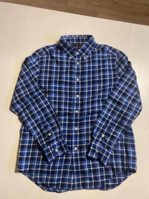 Blårutig skjorta från Ralph Lauren - Klassisk blårutig skjorta från Ralph Lauren med button-down krage och vita knappar. Skjortan har lång ärm och ikonisk broderad logga på bröstet. Tillverkad i mjuk bomull som känns skön mot huden. Perfekt för en stilren och avslappnad look. Skjortan är i storlek M för barn.