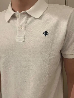 Morris piké - Säljer en stilren vit pikétröja med korta ärmar och krage. Tröjan har knappar framtill och en mörkblå broderad detalj på bröstet. Tillverkad i bomull för en skön och luftig känsla. Perfekt för dig som gillar klassisk och enkel stil.