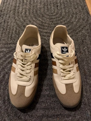 Adidas beige och bruna sneakers - Snygga Adidas sneakers i beige och brunt med klassiska tre ränder på sidan. Ovandelen är i en mix av mocka och syntetmaterial, med vita snören och svart logga på plösen. Perfekta för dig som gillar retrostil och vill ha något som sticker ut.