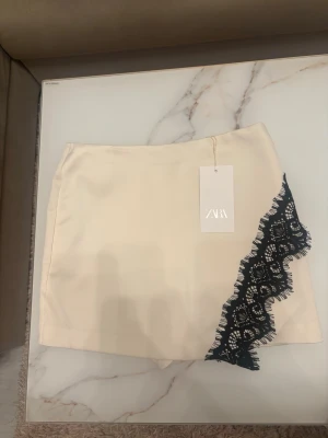 Krämvit byx/kjol med spets från ZARA - Supersnygg krämvit skort från ZARA med unik svart spetsdetalj längs ena sidan. Köpte här på Plick i nytt skick och med lappen kvar. Har ej kommit till användning en ända gång men lappen är borta🥰 