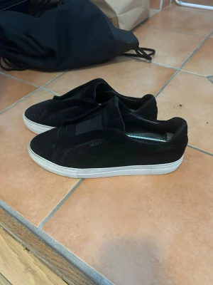 Axel arigato laceless  - Säljer ett par svarta slip-on sneakers i mocka med vit sula. Skorna har en stilren och minimalistisk design utan snörning, vilket gör dem enkla att ta på. Passar perfekt till streetwear och vardagsoutfits.
