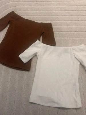 Två ribbade offshoulder-toppar - Säljer två snygga ribbade offshoulder-toppar i vitt och brunt. Båda har en tight passform och korta ärmar, perfekta att styla med jeans eller kjol för en trendig look. Materialet är mjukt och stretchigt, vilket gör dem bekväma att bära hela dagen.