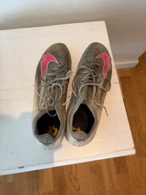 Gråa fotbollsskor med rosa detaljer - Säljer ett par gråa fotbollsskor med snörning och tydliga rosa och gröna detaljer på sidorna. Skorna har en låg profil och är designade för bra bollkontroll på planen. Perfekta för dig som vill sticka ut med färg på matchen.