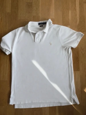 Vit Raffe pikétröja - Klassisk vit pikétröja med korta ärmar och krage. Tröjan har en broderad logga på bröstet och är tillverkad i bomull för en bekväm känsla. Perfekt till jeans eller shorts för en clean look.