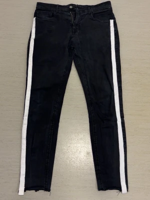 Svarta jeans med vita sidor från Zara - Snygga svarta jeans från Zara Man med raka ben och vita ränder längs sidorna. Jeansen har klassisk femficksmodell, dragkedjegylf och råa benslut för en trendig look. Perfekta för dig som vill sticka ut lite extra.