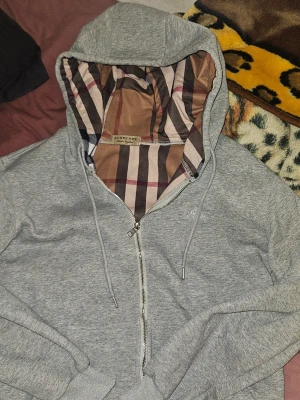 Grå zip hoodie från Burberry - Zipern är lite trög. Snygg grå hoodie med dragkedja från Burberry. Huvan har det klassiska rutmönstret i beige, brunt och rött på insidan. Tröjan har dragsko i huvan och ribbade muddar vid ärmar och nederkant. Perfekt för en chill och stilren look.