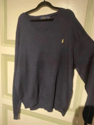 Mörkblå tröja från Polo Ralph Lauren - Mörkblå långärmad tröja från Polo Ralph Lauren med V-ringning och den klassiska gula logotypen broderad på bröstet. Tröjan har ribbade muddar vid ärmslut och nederkant. Perfekt för en avslappnad och stilren look.