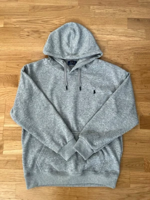 Grå hoodie från Polo Ralph Lauren - Klassisk grå hoodie från Polo Ralph Lauren med huva och dragsnören. Broderad marinblå/svart logga på bröstet och stor känguruficka framtill. Tillverkad i mjuk bomullsblandning och har ribbade muddar vid ärmslut och nederkant. Perfekt för en avslappnad och stilren look. Skriv gärna om ni har några frågor mm, pris kan diskuteras vid rimliga bud.