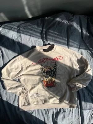 Beige Ed Hardy sweatshirt med tryck - Säljer en beige sweatshirt från Ed Hardy med stort grafiskt tryck framtill som visar en dödskalle med eldflammor och logga i rött. Tröjan har rund hals, långa ärmar och ribbade muddar. Perfekt för dig som gillar streetwear och statementplagg.