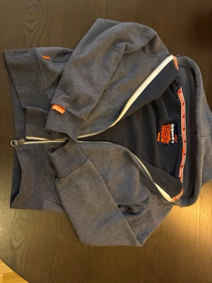 Mörkblå Superdry zip-hoodie - Snygg mörkblå zip-hoodie från Superdry med huva och dragkedja. Orange detaljer och logga på insidan samt vid ärmslut. Mjuk insida och klassisk passform, perfekt för en chill och stilren look.