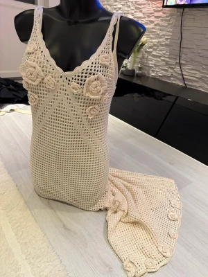 Beige virkad maxiklänning med blommor - Supersnygg beige virkad maxiklänning med smala axelband och dekorativa virkade blommor vid bysten och längs fållen. Klänningen har en figurnära passform och är gjord i ett luftigt, genombrutet material som ger en bohemisk vibe. Perfekt för sommardagar eller festivaler.