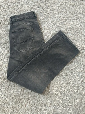 Gråa jeans från Jack & Jones  - Säljer ett par gråa jeans från Jack & Jones  med klassisk femficksmodell och raka ben. Jeansen har en snygg tvättad look och är tillverkade i bomull med lite stretch för extra komfort. Perfekta till en avslappnad stil. 900kr nypris!