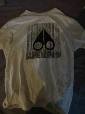 Vit t-shirt med grafiskt tryck - Vit t-shirt med ett stort grafiskt tryck på ryggen som föreställer en svart symbol mot en bakgrund av träd. T-shirten har rund hals och korta ärmar. Materialet ser ut att vara bomull, vilket gör den bekväm och lätt att bära.