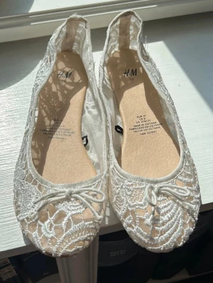 Vita spetsballerinaskor från H&M - Säljer ett par vita ballerinaskor från H&M med vacker spets och små rosetter framtill. Skorna har rund tå och transparent spetsmaterial som ger en romantisk känsla. Perfekta för dig som gillar en feminin och söt stil.
