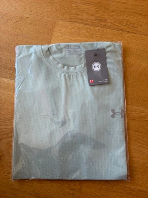 Ljusgrön t-shirt från Under Armour - Snygg ljusgrön t-shirt från Under Armour med diskret logga på bröstet. Tillverkad i ett lätt och ventilerande syntetmaterial som passar perfekt till träning. Klassisk rund hals och korta ärmar för en clean look.
