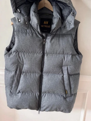 Hetrego väst   - Snygg grå dunväst från Hetrego med avtagbar huva och dragkedja framtill. Västen har två fickor med dragkedja och quiltad design som ger en puffig look. Perfekt för lager-på-lager och håller dig varm under kyliga dagar.
